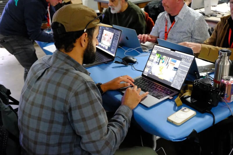 A man in a plaid shirt and baseball cap works on a laptop at a table with other people in a workspace.