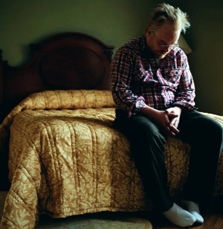 A man with messy blonde hair and glasses sits on the edge of a bed, wearing a plaid shirt and dark pants, with his hands clasped in his lap.