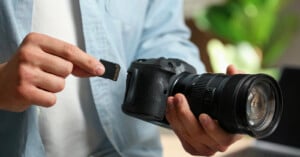 A person in a light blue shirt holds a black camera in one hand and an SD memory card in the other.