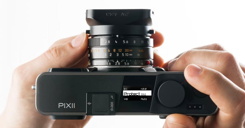 A pair of hands holds a black digital camera from a top-down perspective, showing a small screen displaying settings and a lens with aperture and distance markings.