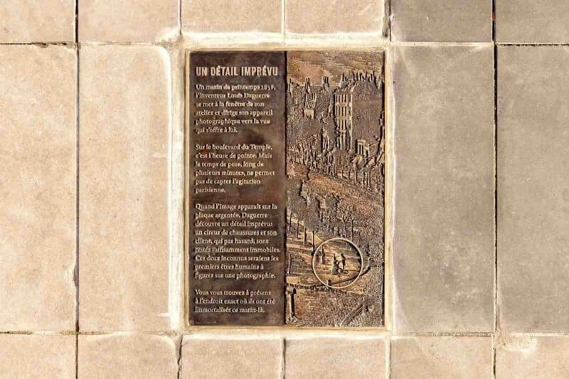 A bronze plaque mounted on a tiled wall features a relief of a historic street scene and French text describing Louis Daguerre's 1838 photograph.