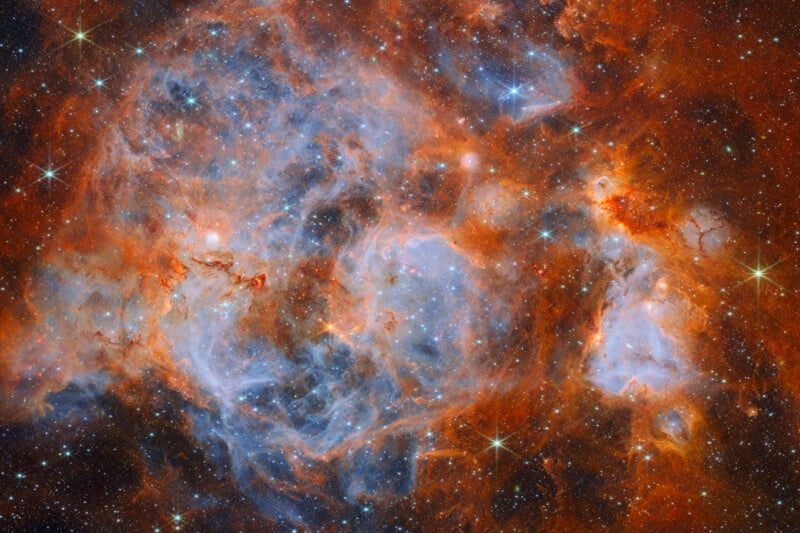 A vibrant nebula filled with glowing orange gas clouds, swirling blue dust, and numerous bright, sparkling stars scattered throughout the frame.