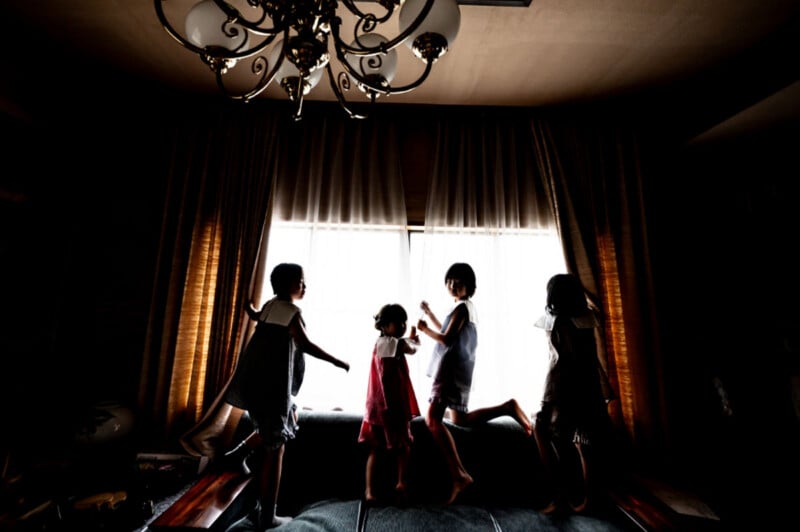 Four children stand in silhouette on a sofa in front of a bright window, with a chandelier hanging above them.