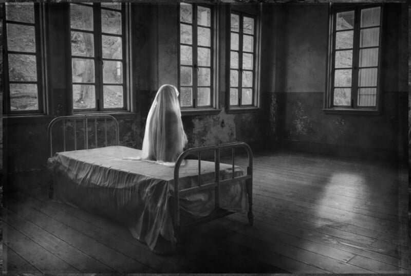 A figure draped in a white sheet sits on a metal-framed bed in a dimly lit, empty room with large windows.