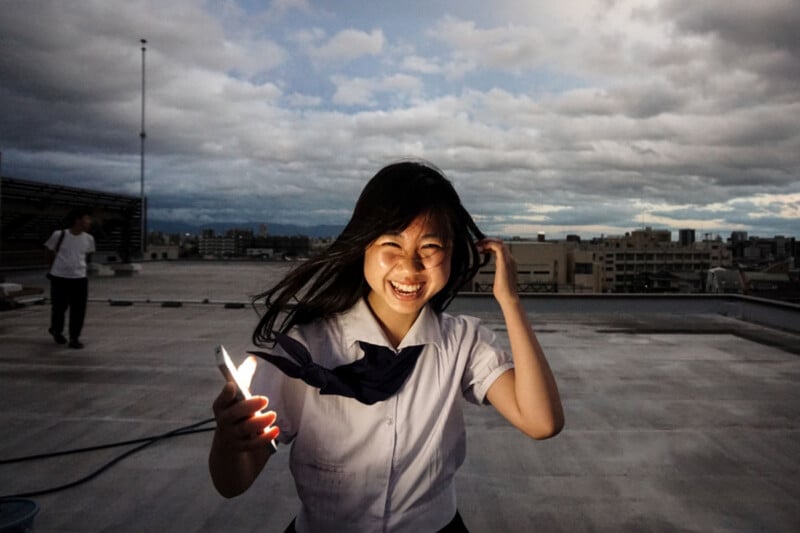 A smiling girl in a school uniform stands on a rooftop at dusk, holding a glowing smartphone that illuminates her face.