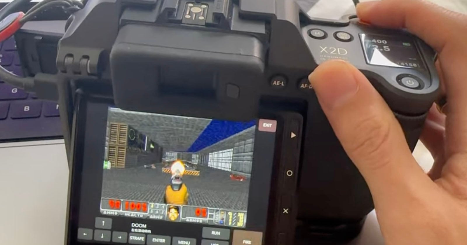 A person's hand holds a digital camera displaying the video game Doom on its rear screen, with a virtual keyboard overlay at the bottom.