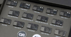 A collection of Ricoh GR camera pins arranged in a grid on a dark gray display board, each labeled with its model name and year.