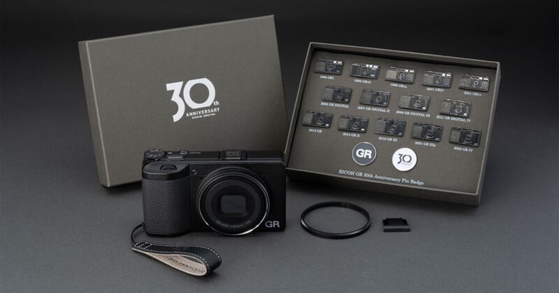 A Ricoh GR 30th Anniversary camera set including a camera with a wrist strap, a lens ring, a hot shoe cover, and a display box containing 15 miniature camera pin badges.