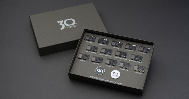 A Ricoh GR 30th Anniversary pin badge set in a black box, featuring miniature metal pins of various camera models and two circular logo pins.