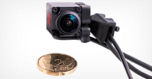 A small, black, cube-shaped camera with a wide-angle lens sits next to a 20 Euro cent coin on a white background.