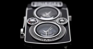 A close-up of a black and silver watch designed to resemble a vintage twin-lens reflex camera, featuring two circular clock faces and the text "FRANKE & HEIDECKE GERMANY" on the lower lens area.