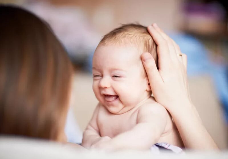 A person gently holds the head of a smiling baby who is looking toward them.