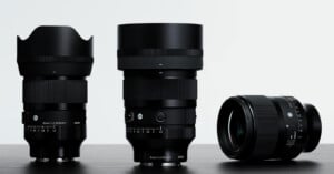 Three black Sigma camera lenses stand on a dark surface against a white background, with two standing upright and one lying on its side.