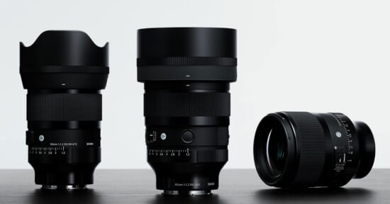 Three black Sigma camera lenses stand on a dark surface against a white background, with two standing upright and one lying on its side.