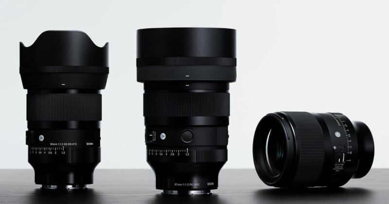 Three black Sigma camera lenses stand on a dark surface against a white background, with two standing upright and one lying on its side.