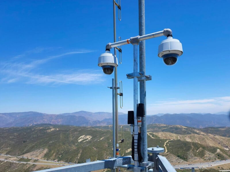 Two dome security cameras are mounted on a metal pole overlooking a vast, mountainous landscape with a highway below.