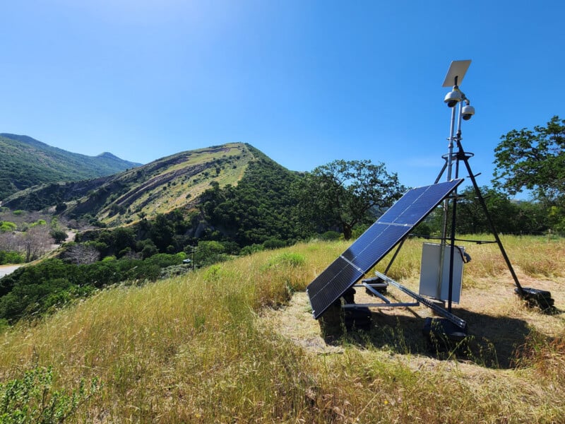 A solar-powered remote monitoring station with cameras and an antenna stands on a grassy hillside overlooking a mountain range.