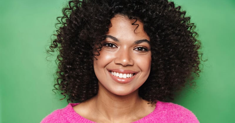 A smiling woman with dark, curly hair wears a bright pink sweater against a solid green background.