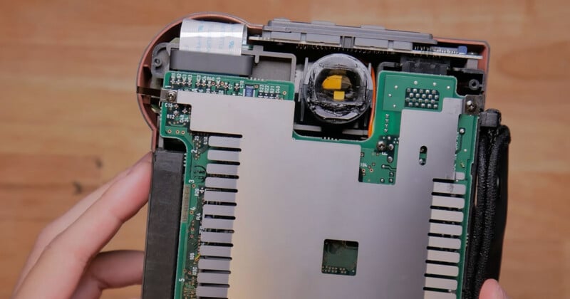 A close-up view of the internal components of an electronic device, showing a green circuit board partially covered by a metal heat sink and a circular lens assembly.