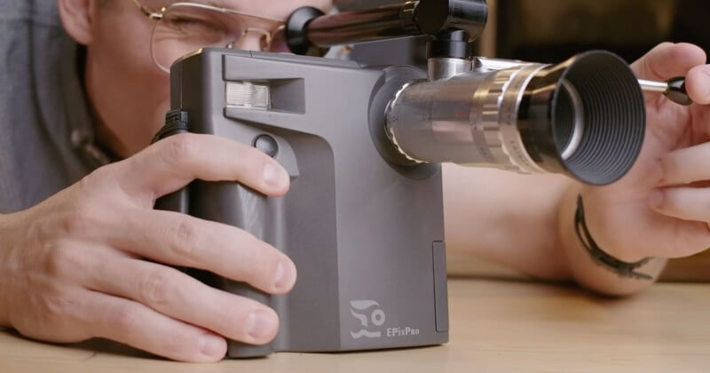 A person looks through the viewfinder of a gray EPixPro camera while holding the lens with their right hand.