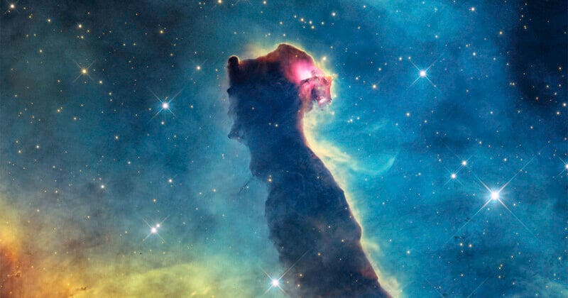 A dark, towering pillar of interstellar gas and dust stands against a vibrant blue nebula filled with scattered bright stars.