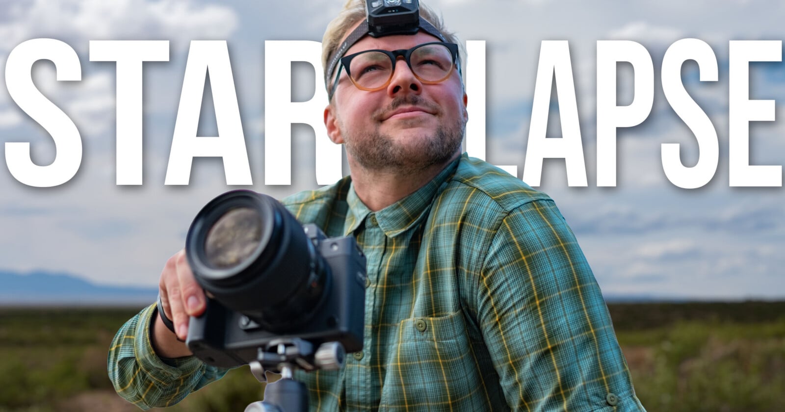 A man wearing glasses and a headlamp looks upward while holding a camera mounted on a tripod against a cloudy sky background.