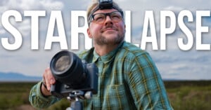 A man wearing glasses and a headlamp looks upward while holding a camera mounted on a tripod against a cloudy sky background.