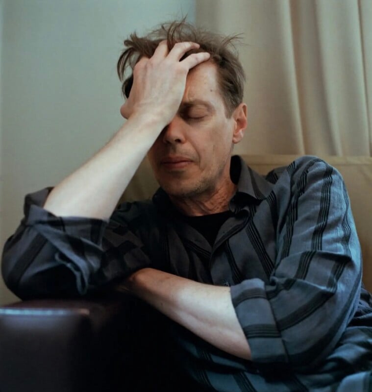 A man with messy hair sits with his eyes closed, resting his forehead against his hand in a gesture of exhaustion.