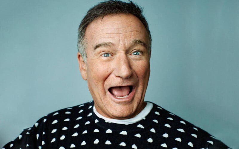 Robin Williams smiles widely with his mouth open against a plain blue background, wearing a black sweater with a white cloud pattern.