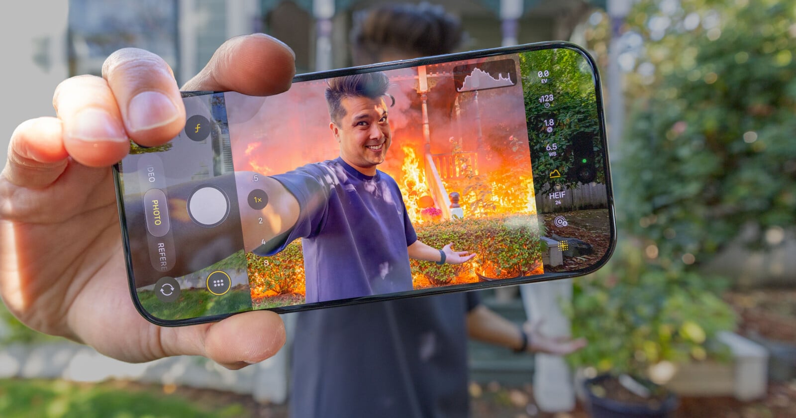 A hand holds a smartphone displaying a camera interface, showing a man smiling in front of a house engulfed in flames.