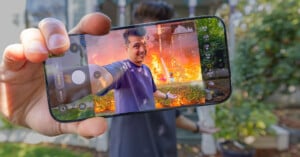 A hand holds a smartphone displaying a camera interface, showing a man smiling in front of a house engulfed in flames.