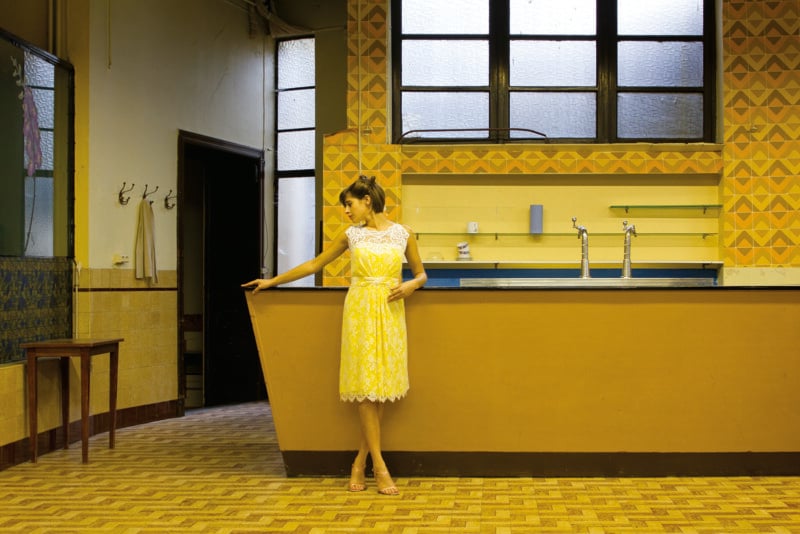 A woman in a yellow lace dress stands leaning against a bar counter in a room with yellow patterned walls and tiled floors.