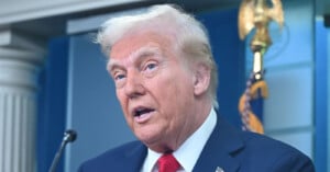 Donald Trump speaks at a podium with a blue background and a gold eagle emblem visible behind him.
