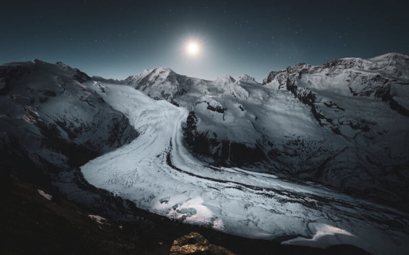 A bright moon shines over a vast, snow-covered glacier nestled between dark, rugged mountain peaks under a starry night sky.