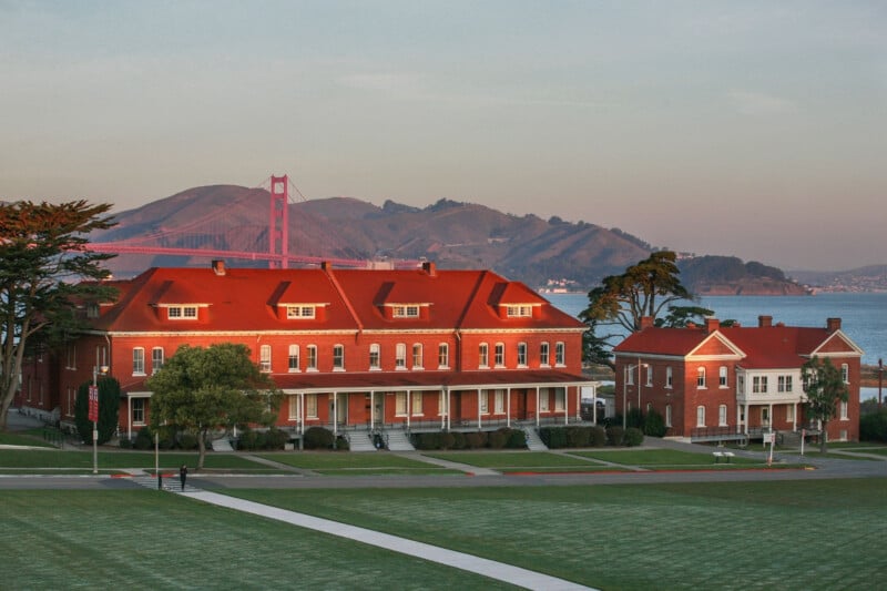 Two red-brick buildings with red roofs sit on a grassy lawn with the Golden Gate Bridge visible in the background.