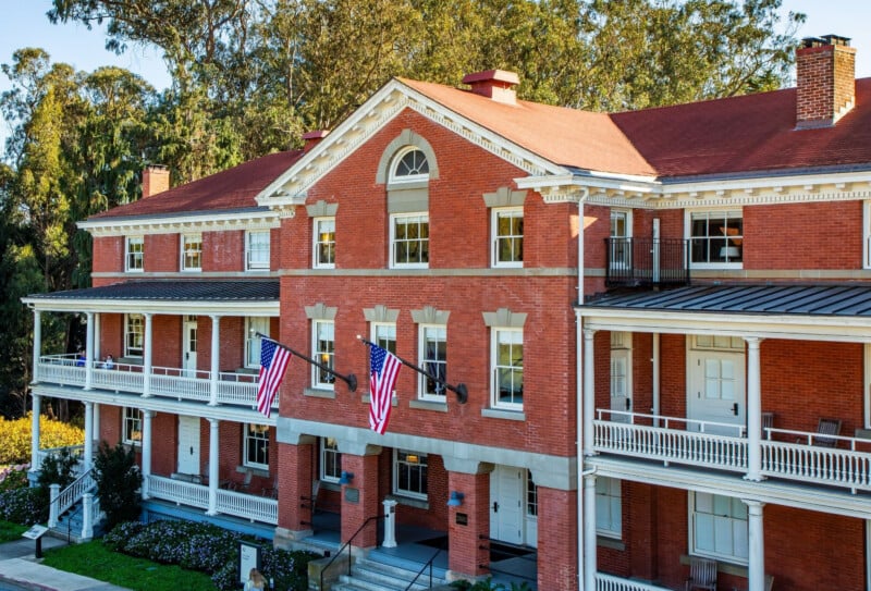 A three-story red brick building with a gabled roof, white trim, and two-level porches, featuring two American flags mounted on the front facade.