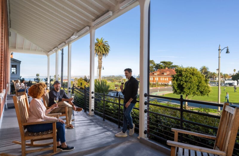 Three people sit and stand on a wooden porch with rocking chairs, holding wine glasses and looking out over a park and a distant bay.