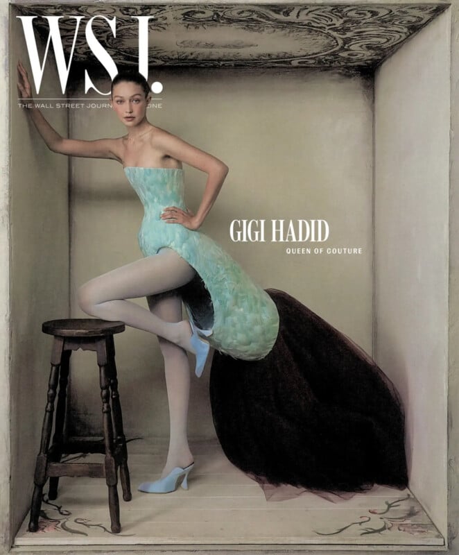 Gigi Hadid poses in a structured, light blue feathered dress with a dark train, leaning against a wall next to a wooden stool.