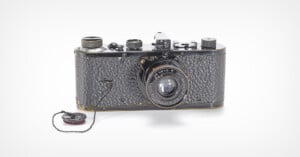 A vintage black film camera with a textured body and a lens labeled "Leitz Anastigmat 1:3,5 F=50mm," featuring a lens cap attached by a string.