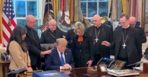 Donald Trump sits at the Resolute Desk with his head bowed, surrounded by a group of clergy members who have their hands on his shoulders and back in prayer.