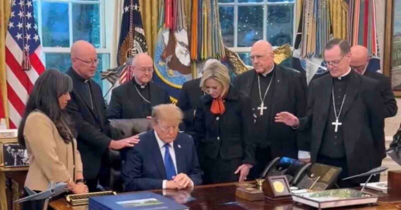 Donald Trump sits at the Resolute Desk with his head bowed, surrounded by a group of clergy members who have their hands on his shoulders and back in prayer.