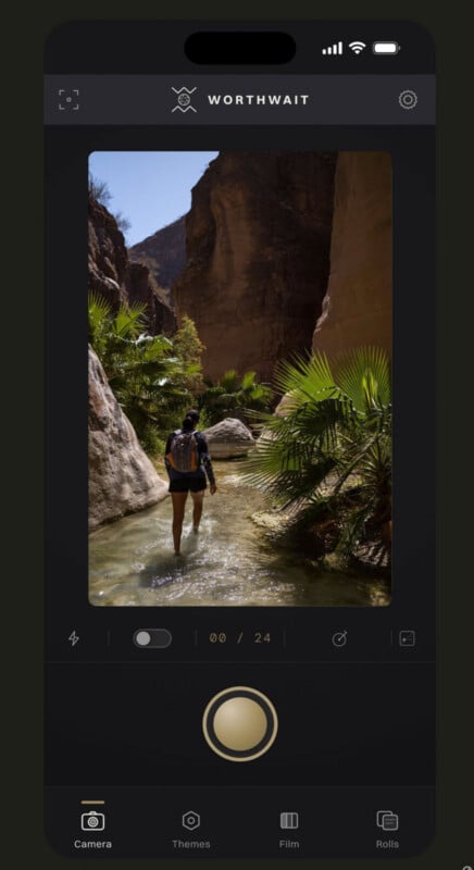 A smartphone screen displays the Worthwait camera app interface, showing a person wading through a shallow stream in a canyon.