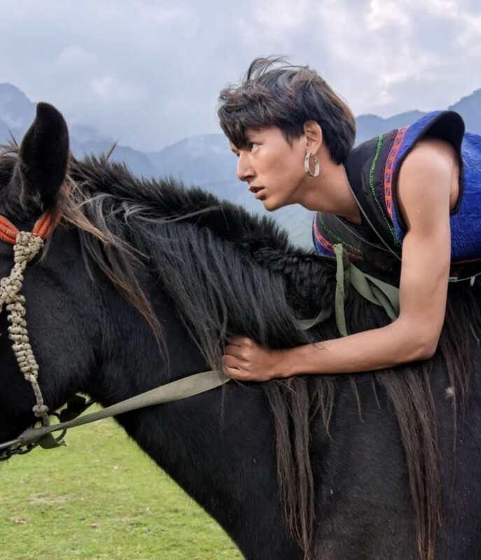 A young person with dark hair and a hoop earring leans forward over the neck of a black horse against a mountain backdrop.