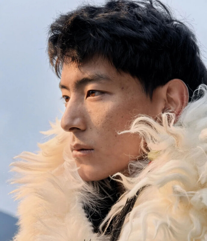 A close-up profile shot of a young man with dark hair and freckles, wearing a thick, white faux-fur collar against a pale sky.