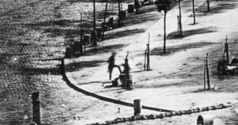 A blurry, black-and-white long-exposure photograph shows a person standing on a sidewalk while getting their shoes shined.