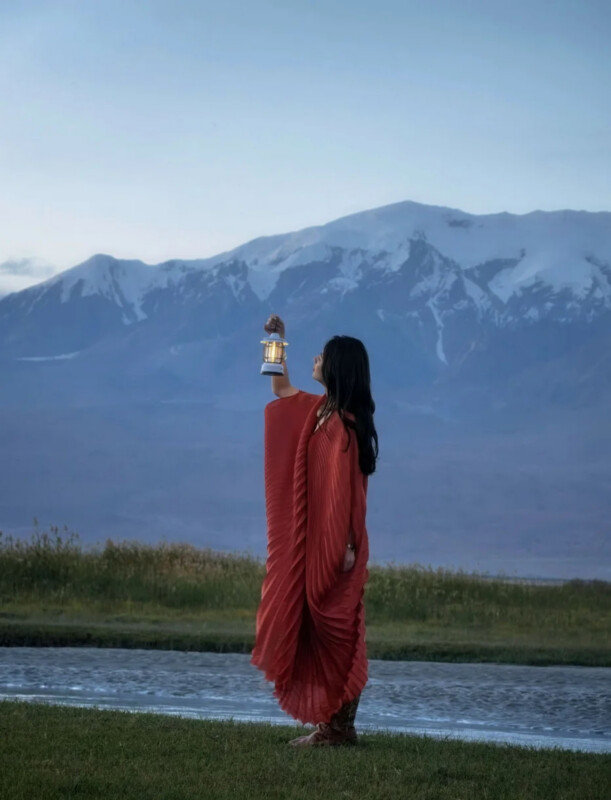 A woman in a long, pleated red dress stands by a river, holding a small glowing lantern up toward a large, snow-capped mountain range.